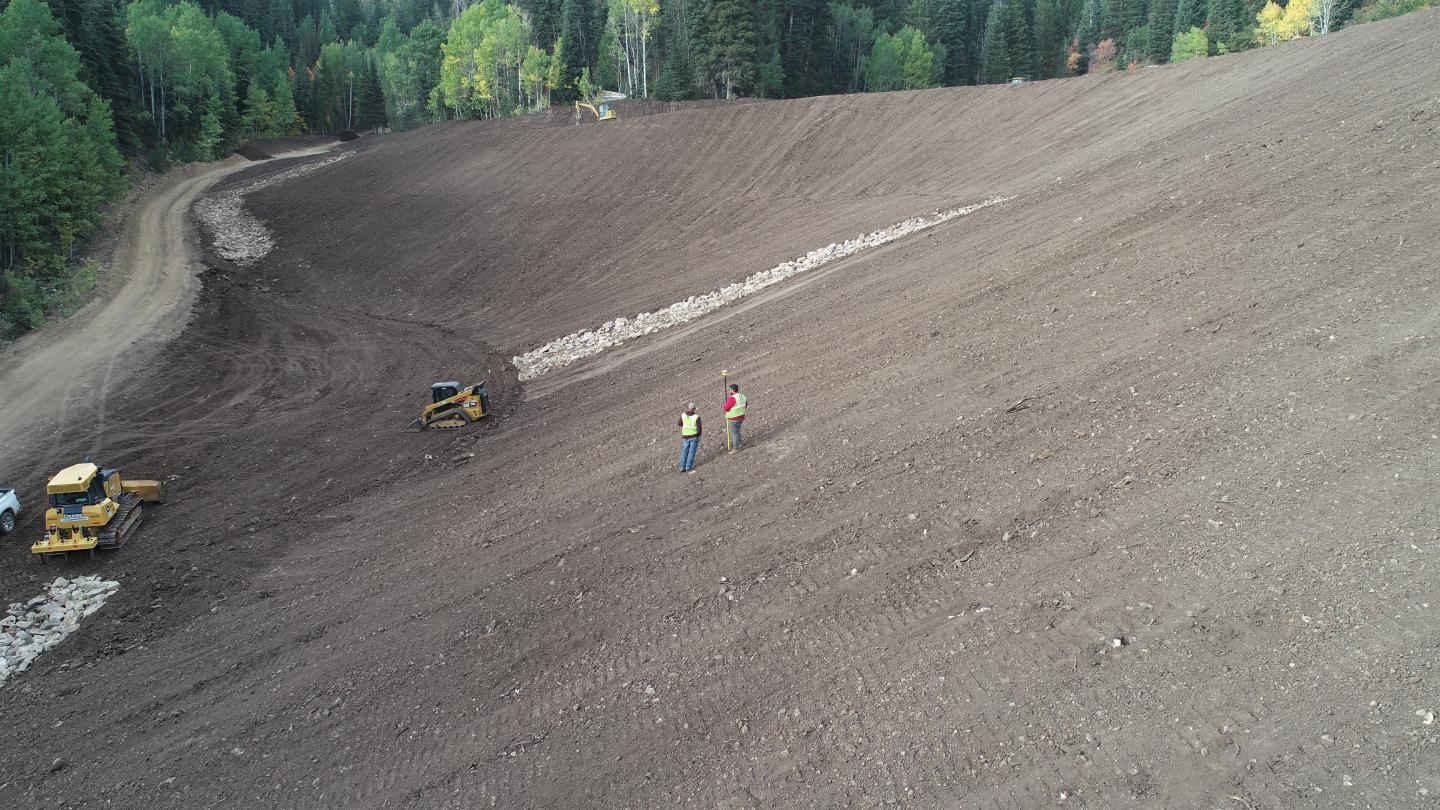 Construction of a ski run at Deer Valley. Two men standing on dirt slope with excavators in background.