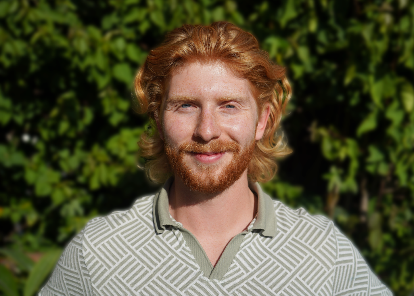 Smiling man with red hair in a green patterned shirt, greenery background.