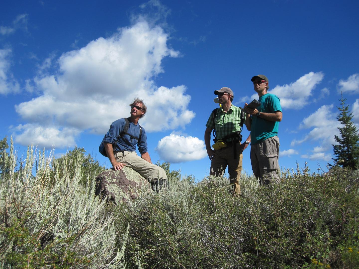Three people standing in a scenic landscape under a blue sky with clouds.