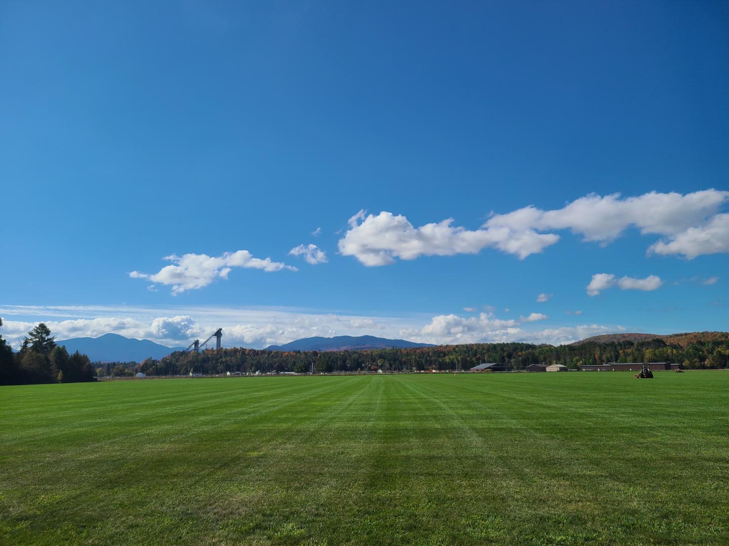 Green field under clear blue sky with clouds and distant mountains.