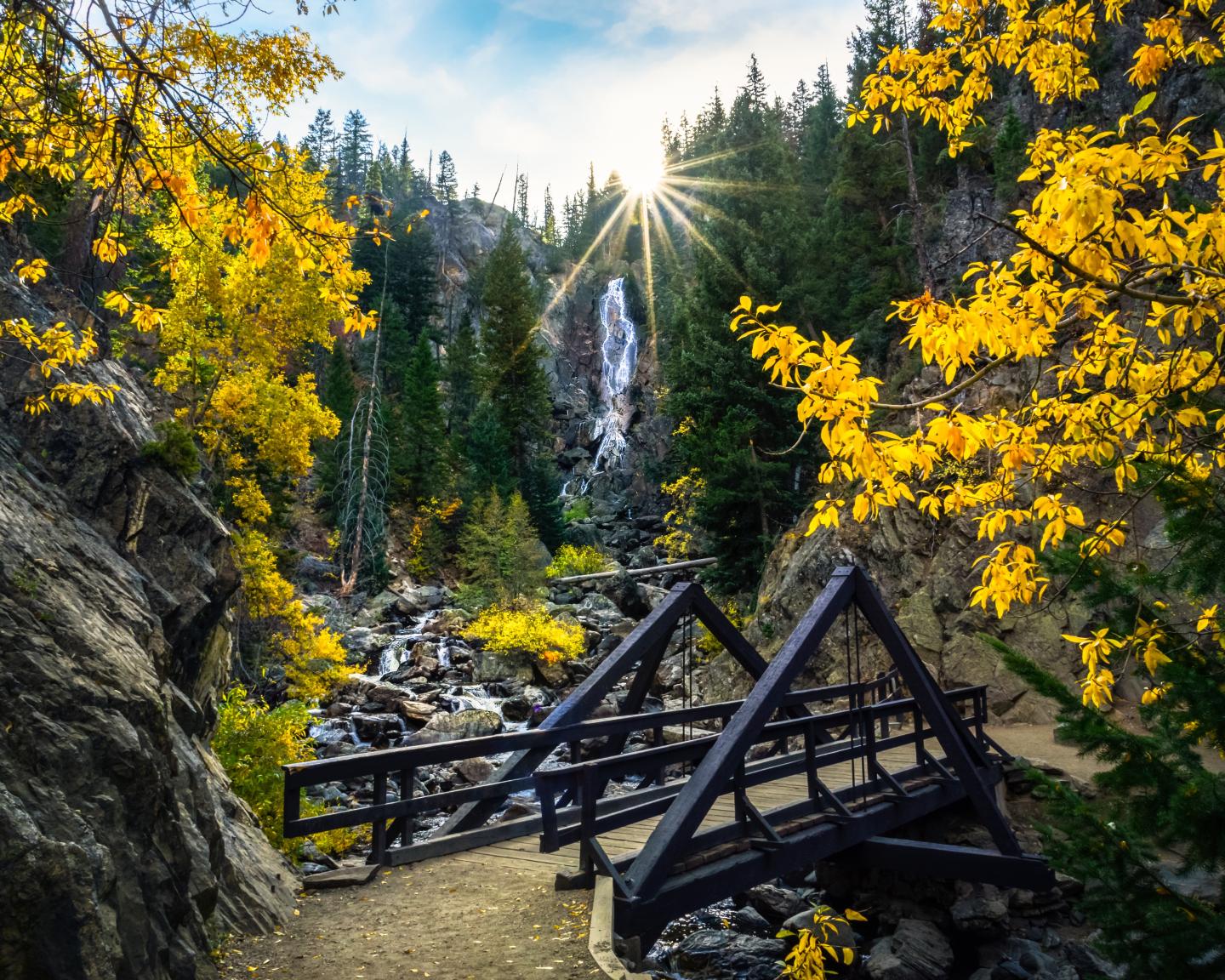 Wooden bridge in forest, surrounded by autumn trees and a waterfall in the background.