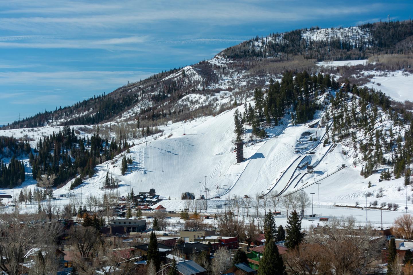 Snowy mountain with ski slopes, trees, and buildings below under a clear blue sky.