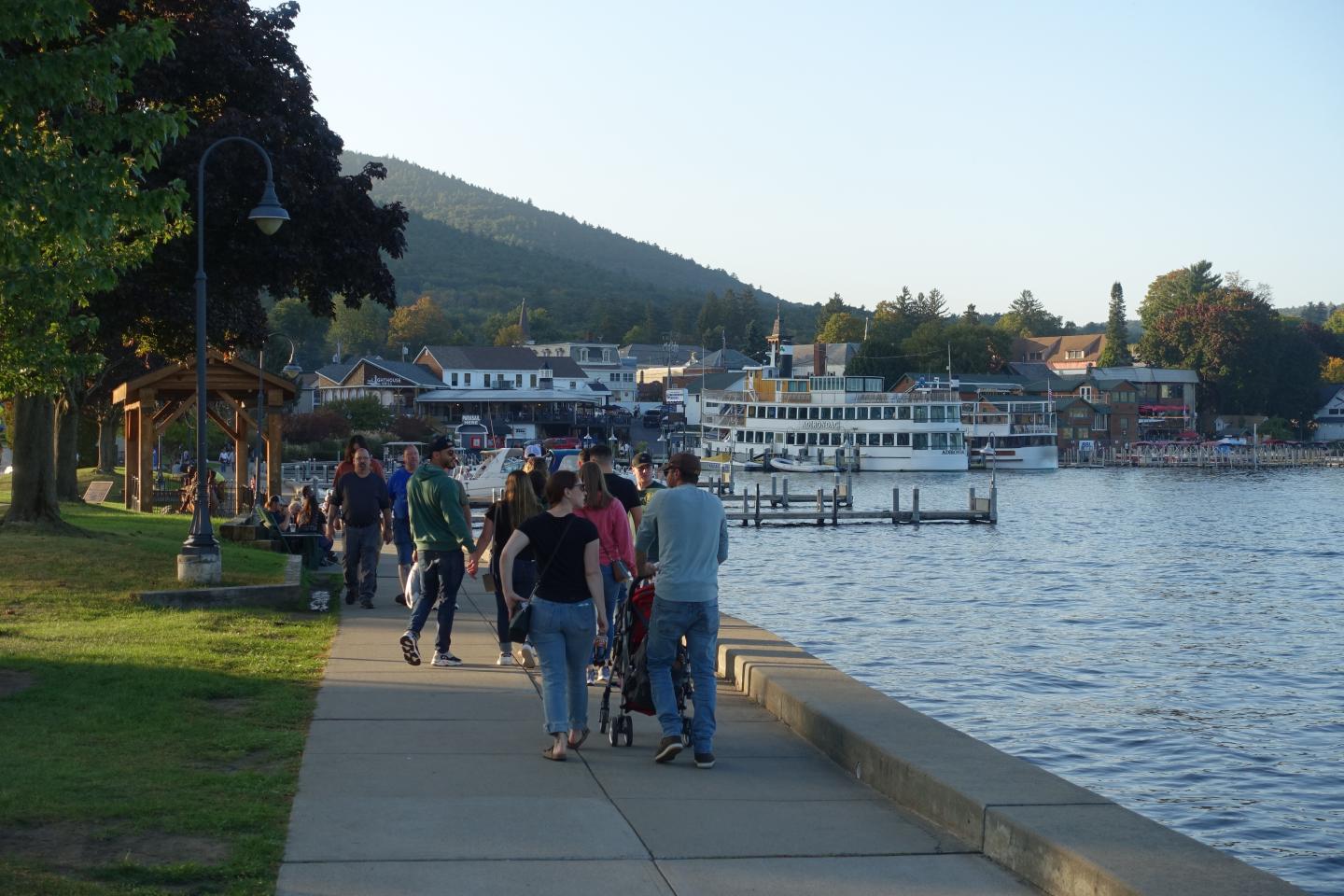 People strolling by a lake, docked boats, distant mountains, and evening light.