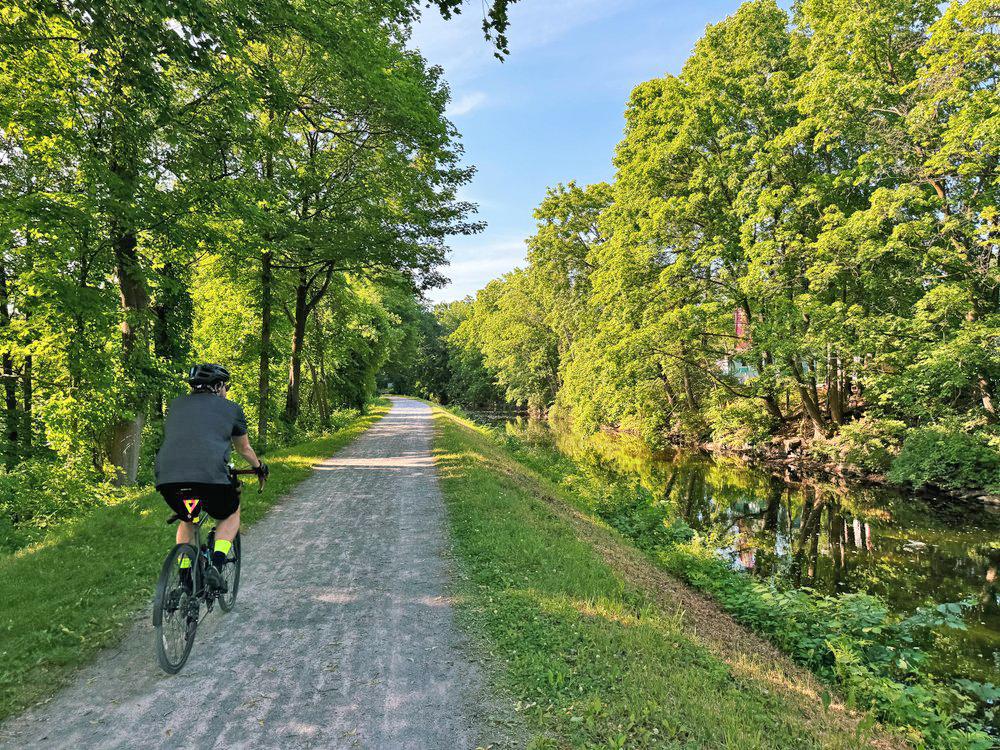 Cyclist on a tree-lined path beside a serene river under a blue sky.