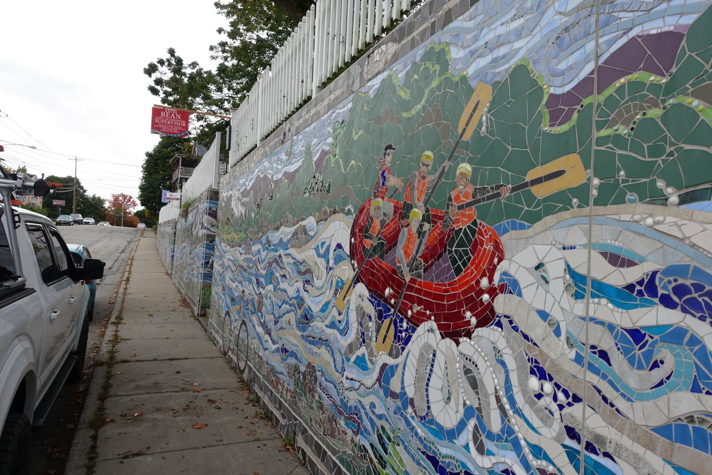 Mosaic of three people rowing a boat on colorful waves along a street.