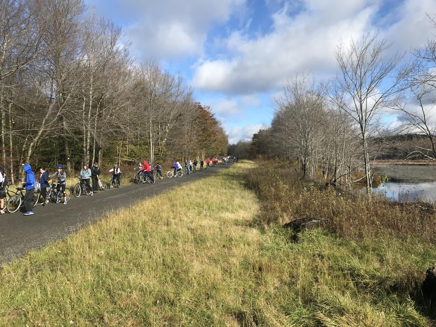 Group walking on a trail beside a lake, under a cloudy sky.