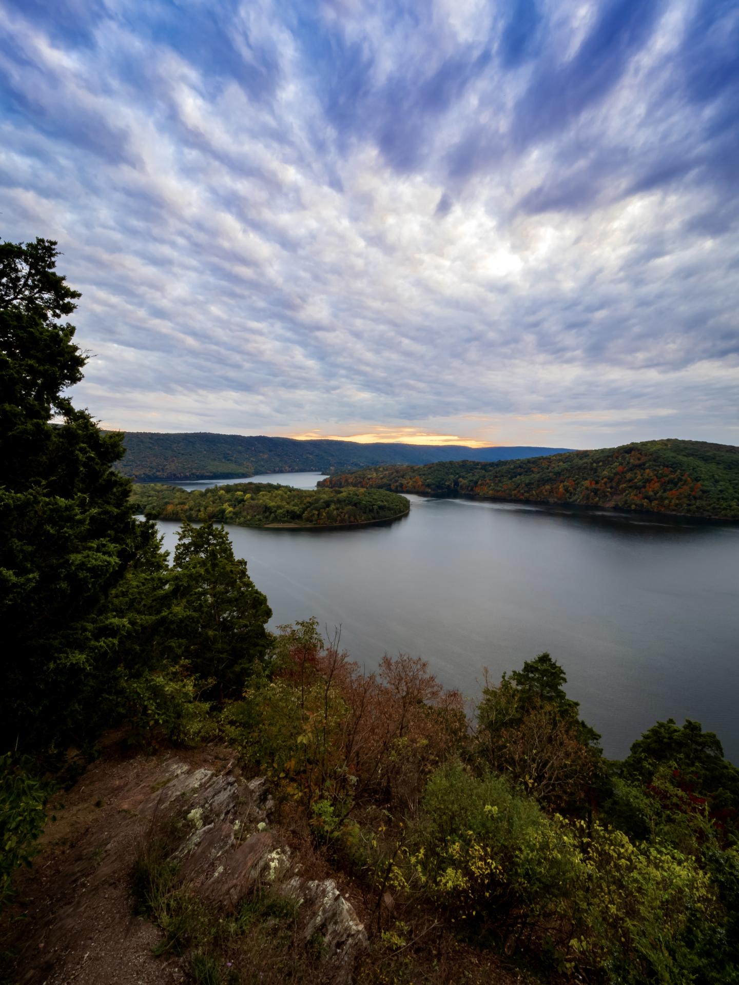 Cloudy sky over a serene lake with surrounding hills and a rocky path in the foreground.