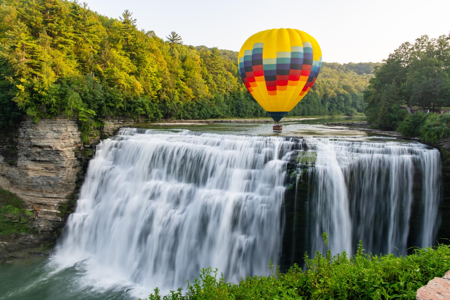 Hot air balloon floating above a waterfall surrounded by lush trees.
