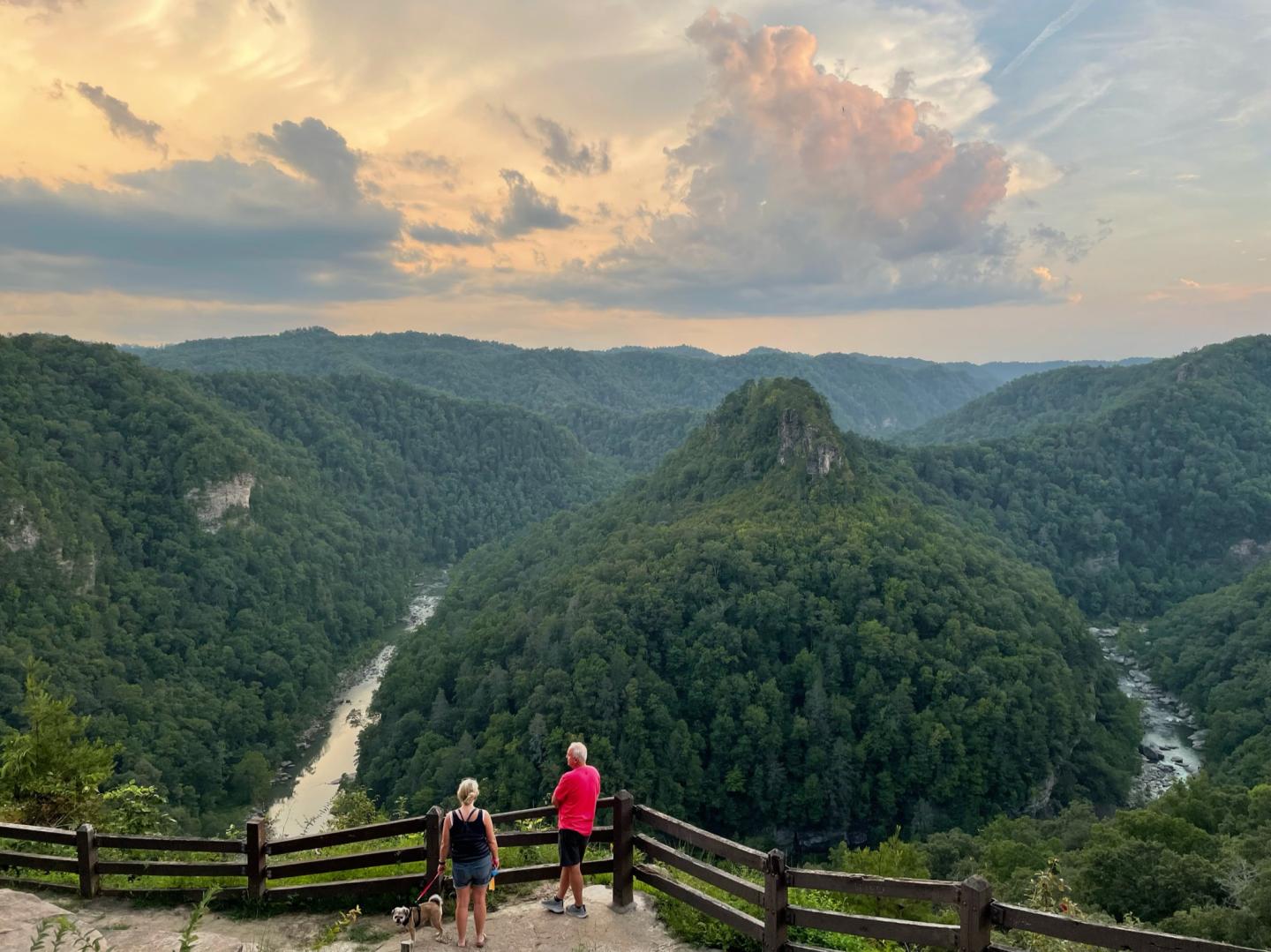 Two people overlook a lush, green valley at sunset with dramatic clouds above.