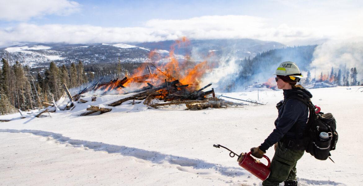 Firefighter with a helmet holds a fire extinguisher near a controlled burn in a snowy landscape.