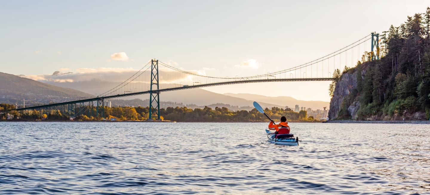 Kayaker paddles near a bridge at sunset with mountains in the background.