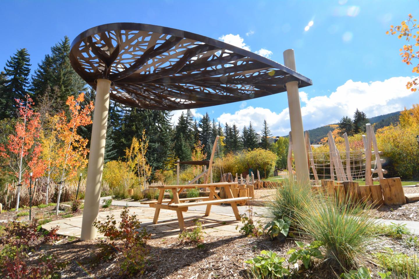 Outdoor park with wooden canopy, picnic table, autumn trees, and blue sky.