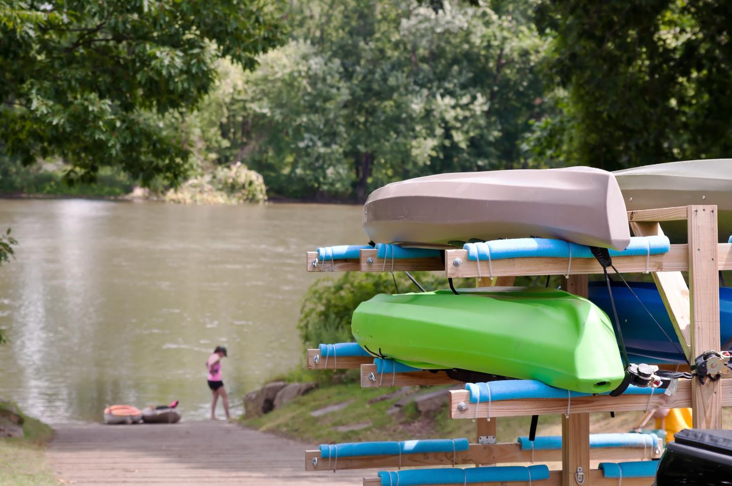 Kayaks stacked near a river with a woman by the water.