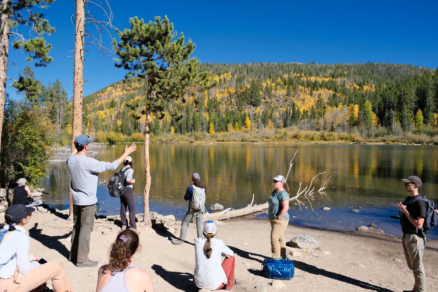 Group of people by a lake, surrounded by trees under a clear blue sky.