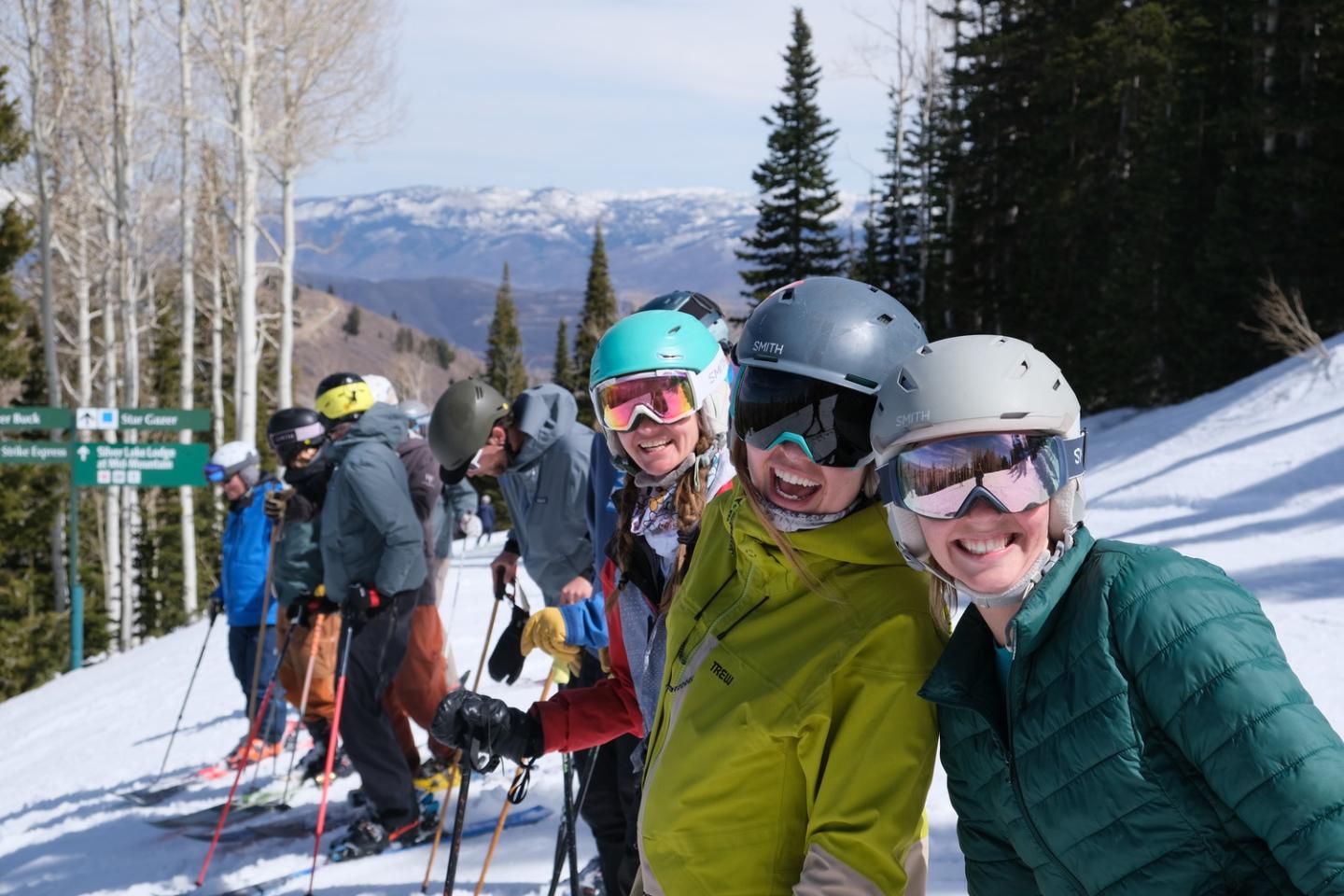 Skiers lined up on a snowy slope, smiling with mountains in the background.