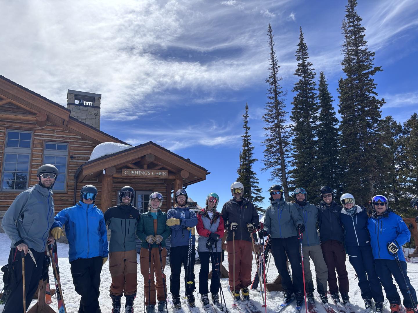 Group of skiers in colorful gear standing in front of a wooden lodge and snowy trees.