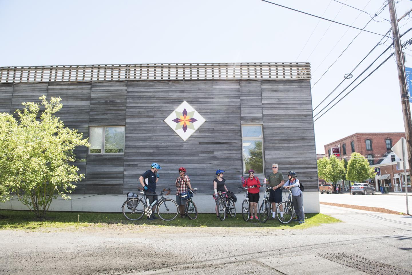 Group of cyclists resting against a wooden building on a sunny day.